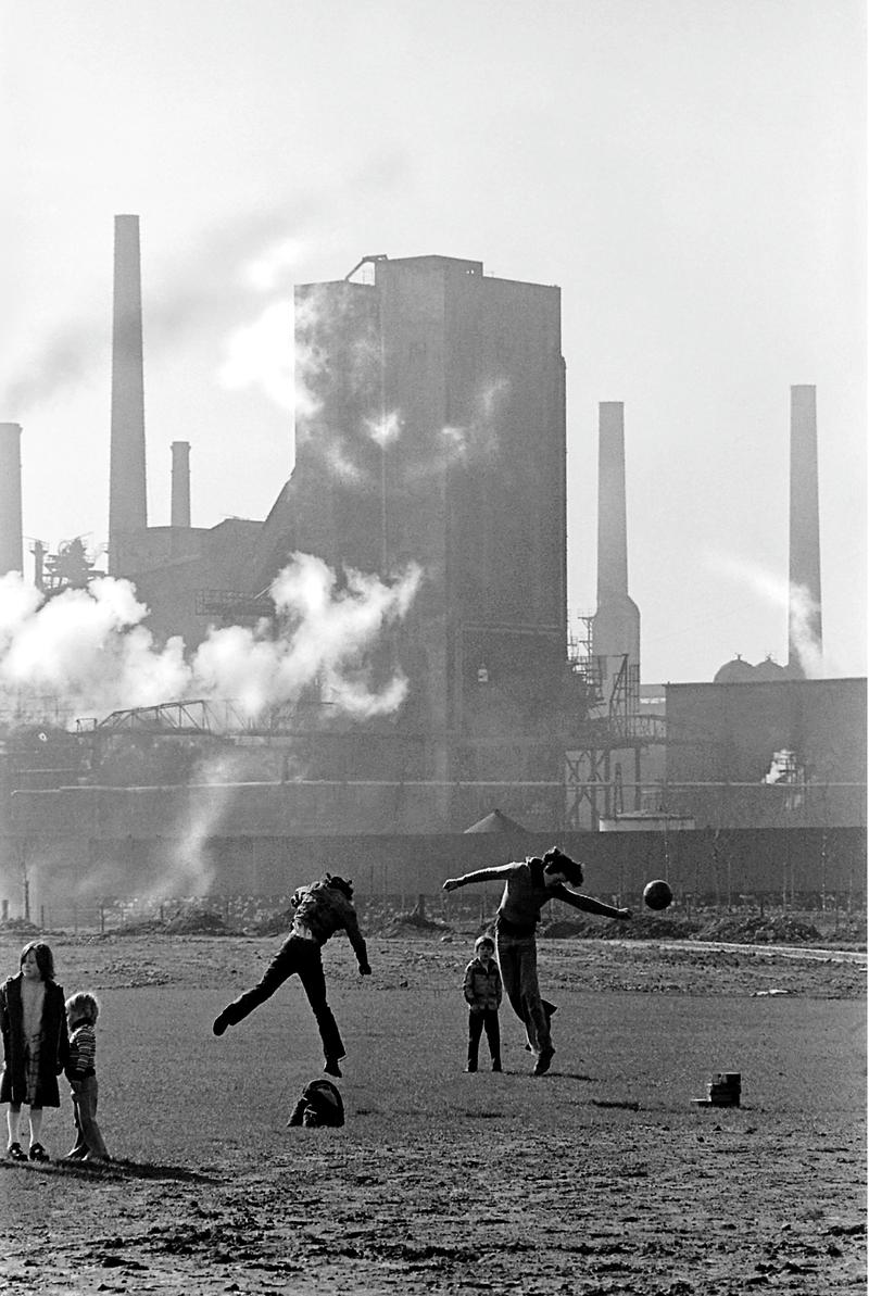 UK. Wales. Cardiff.  Children play football in spare land in front of East Moors steel works at the time that the steel works were closed.  Perhaps their fathers are now out of work.  Cardiff. 1978.