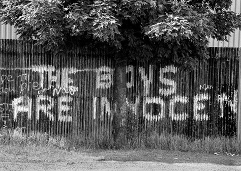Black-and-white photograph of some large graffiti daubed onto a corrugated iron fence. The graffiti reads ‘THE BOYS ARE INNOCENT’ and is partially obscured by a tree.