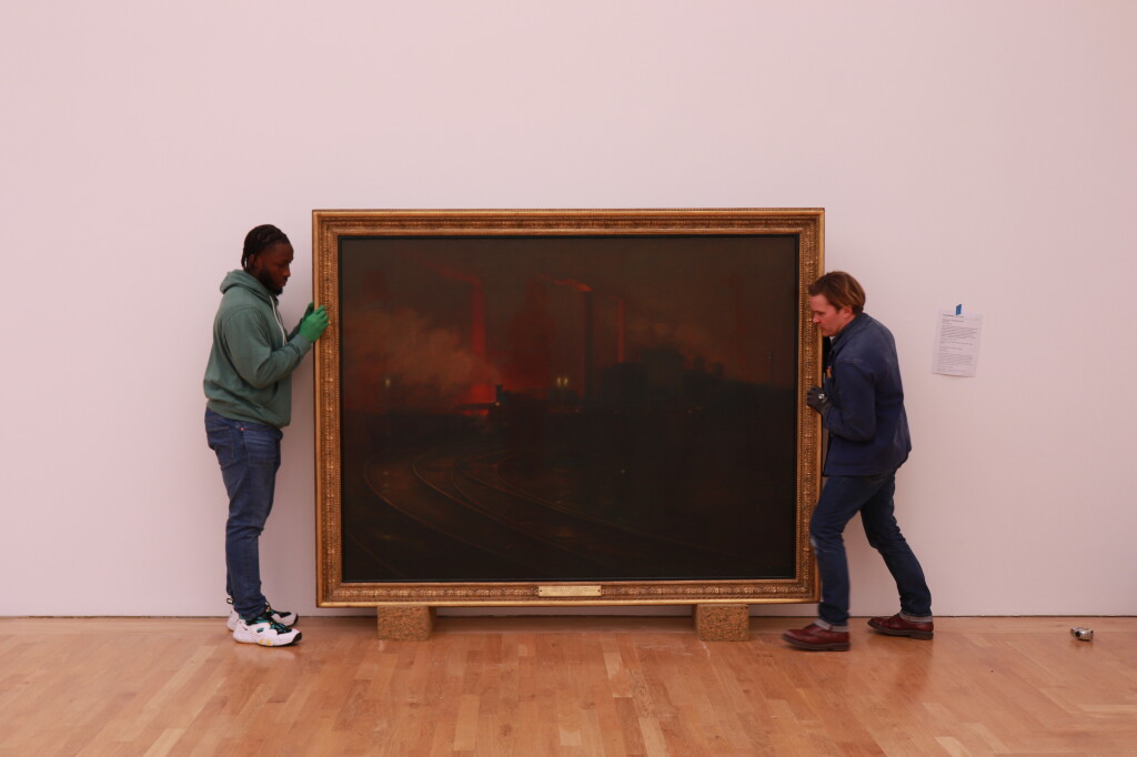 Two people handling a large, landscape-format painting of a dark, industrial scene.