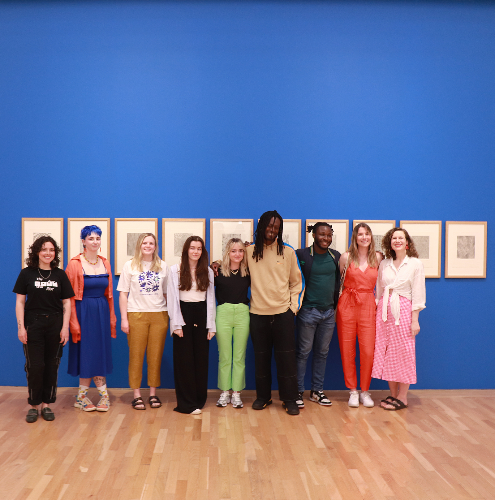 A group of nine people standing in front of a row of prints hanging on a deep blue wall in an art gallery