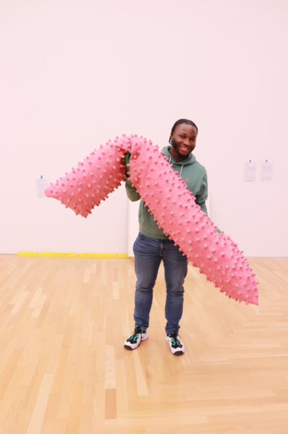 A young man in an art gallery holding a soft, pink, knobbly object about as long as he is tall.