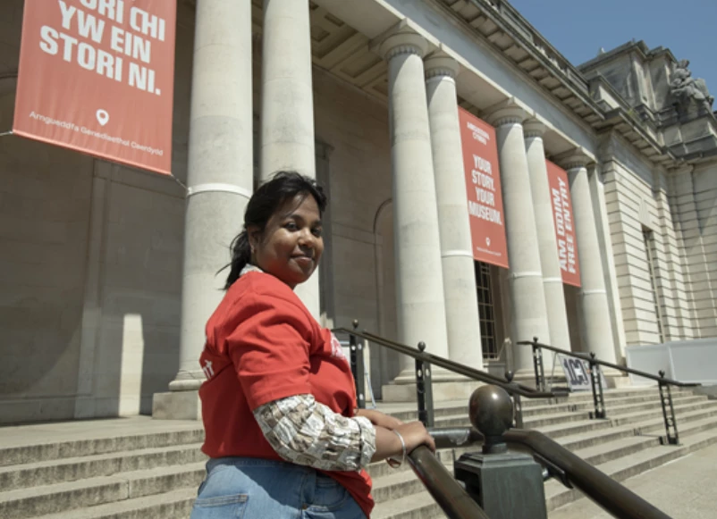 Photograph of a person dressed in red by the steps of a grand building with columns and red banners, under a clear sky.
