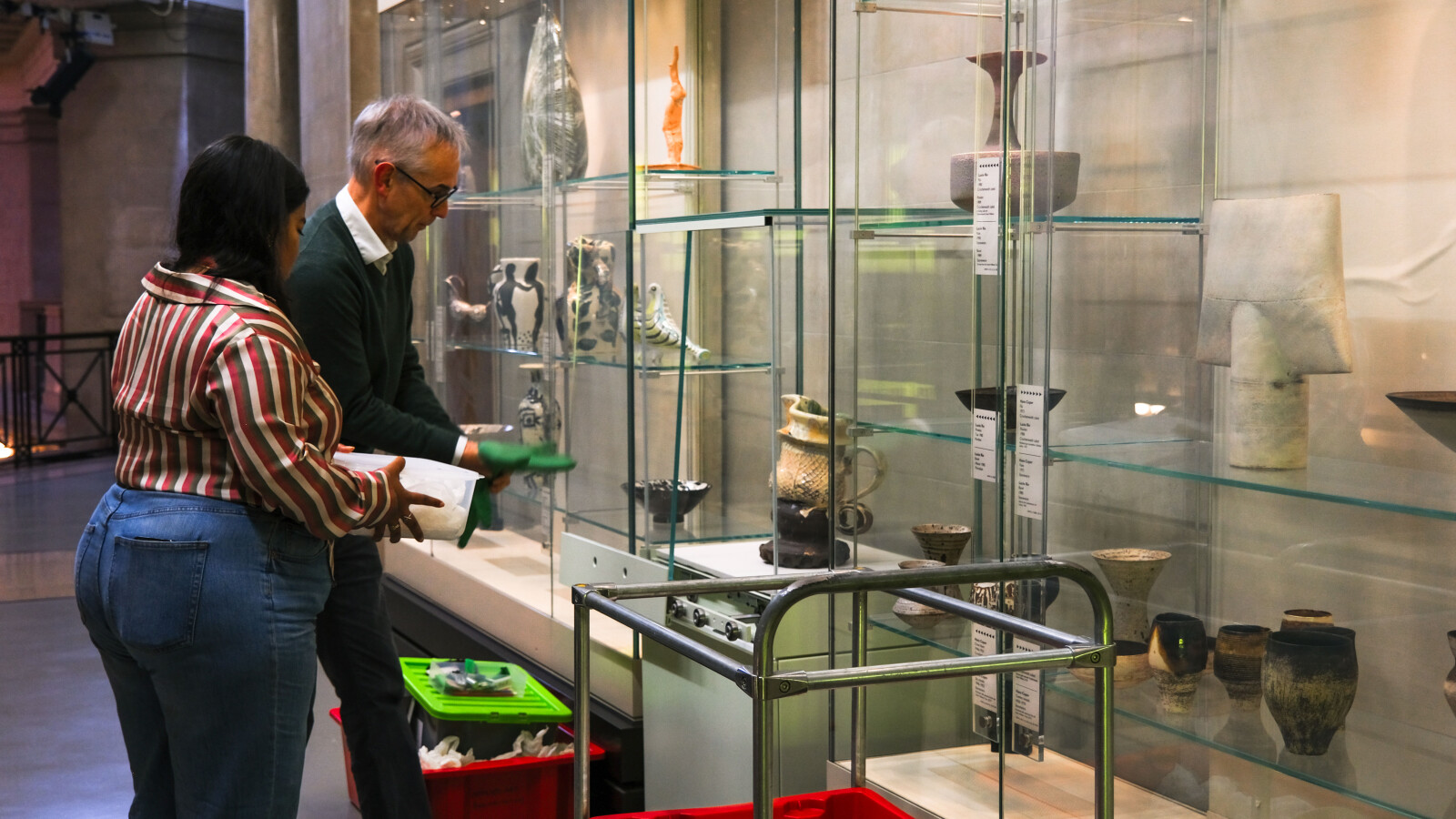 Two people arrange a glass museum case with a variety of ceramic objects.