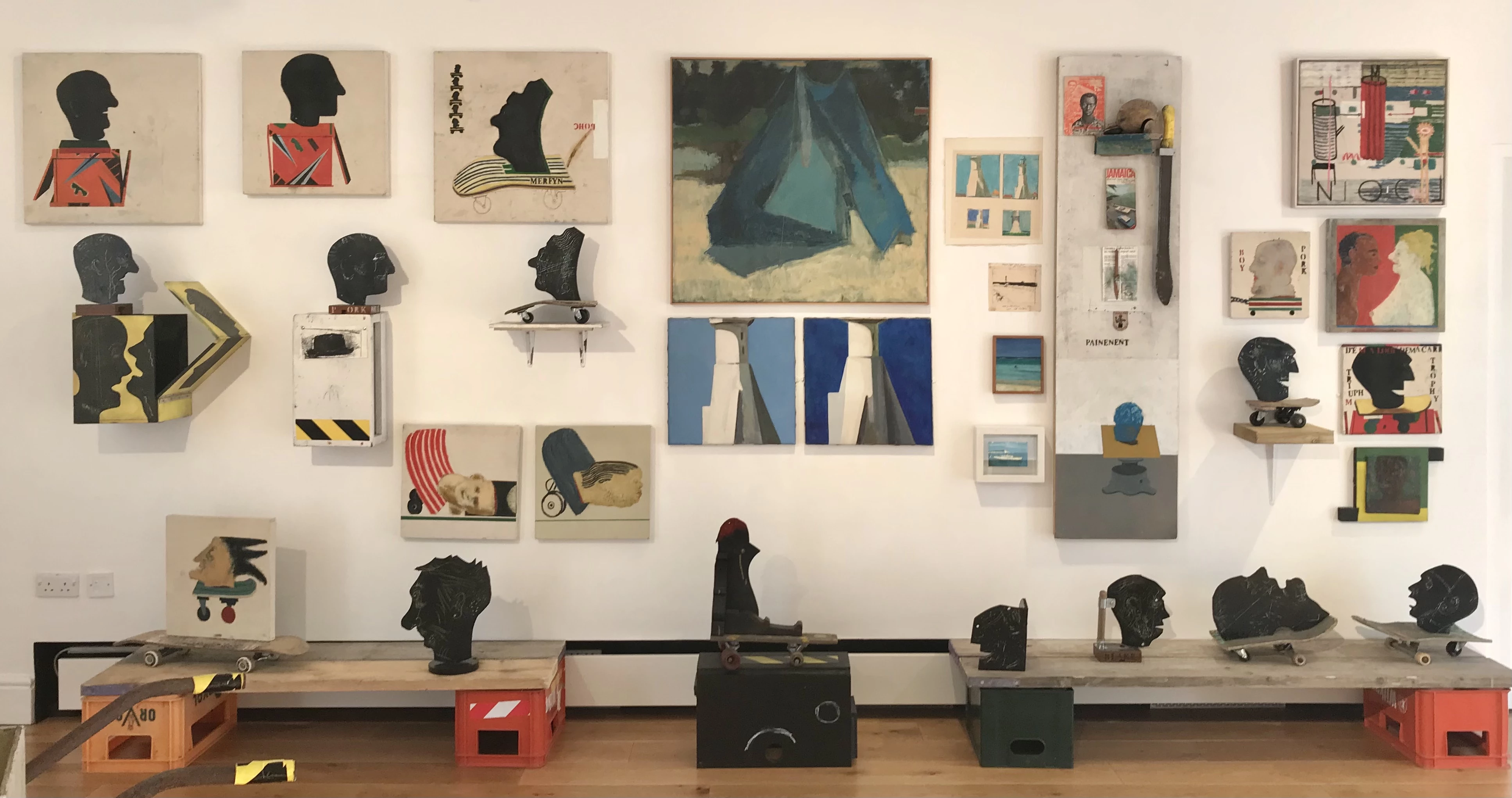 A variety of artworks on and in front of a white wall. The artworks vary from paintings to sculptures showing figures and shapes.