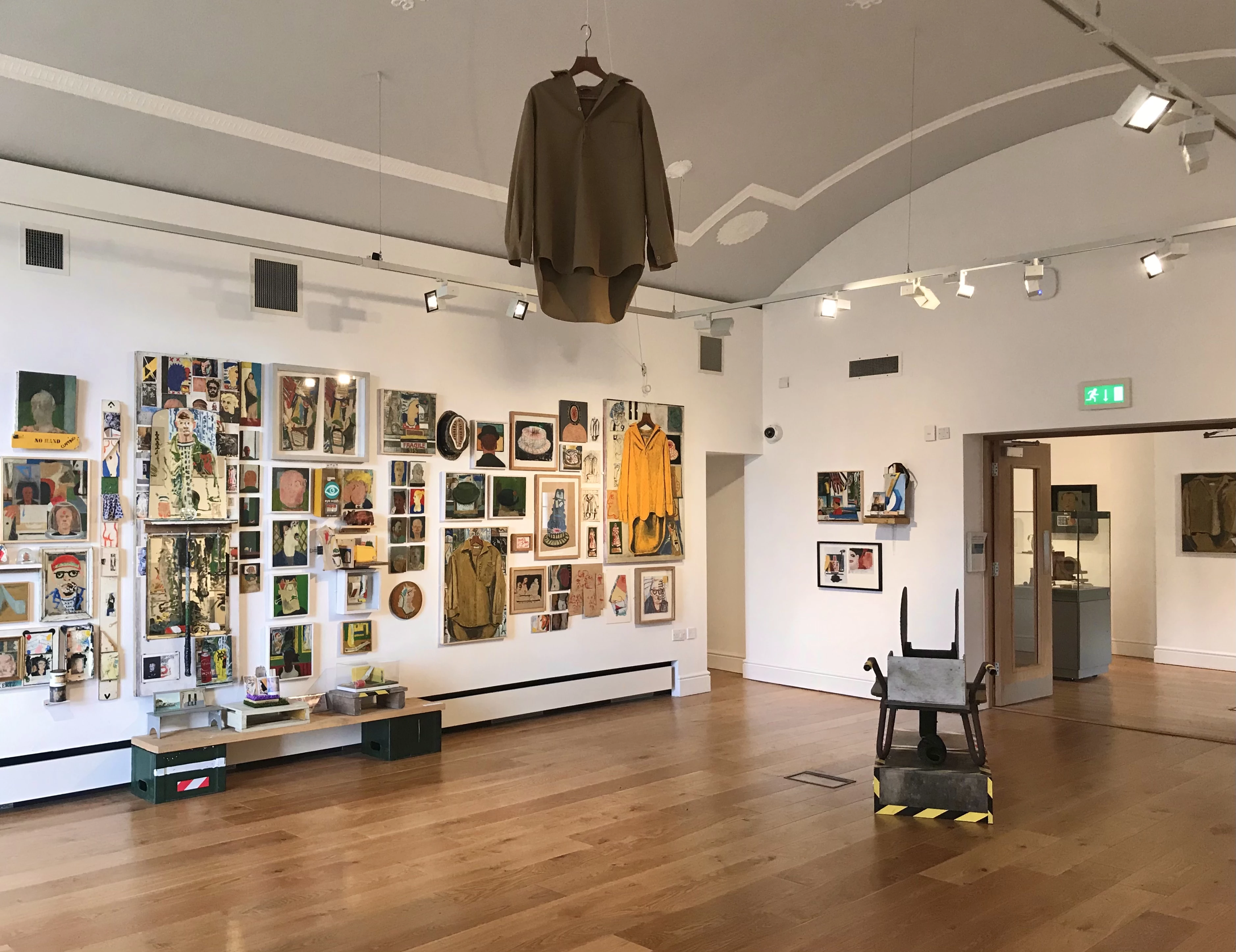 A variety of colourful paintings on a white wall to the left, and a brown shirt hangs in the centre from the ceiling. On the right is a wheelbarrow.