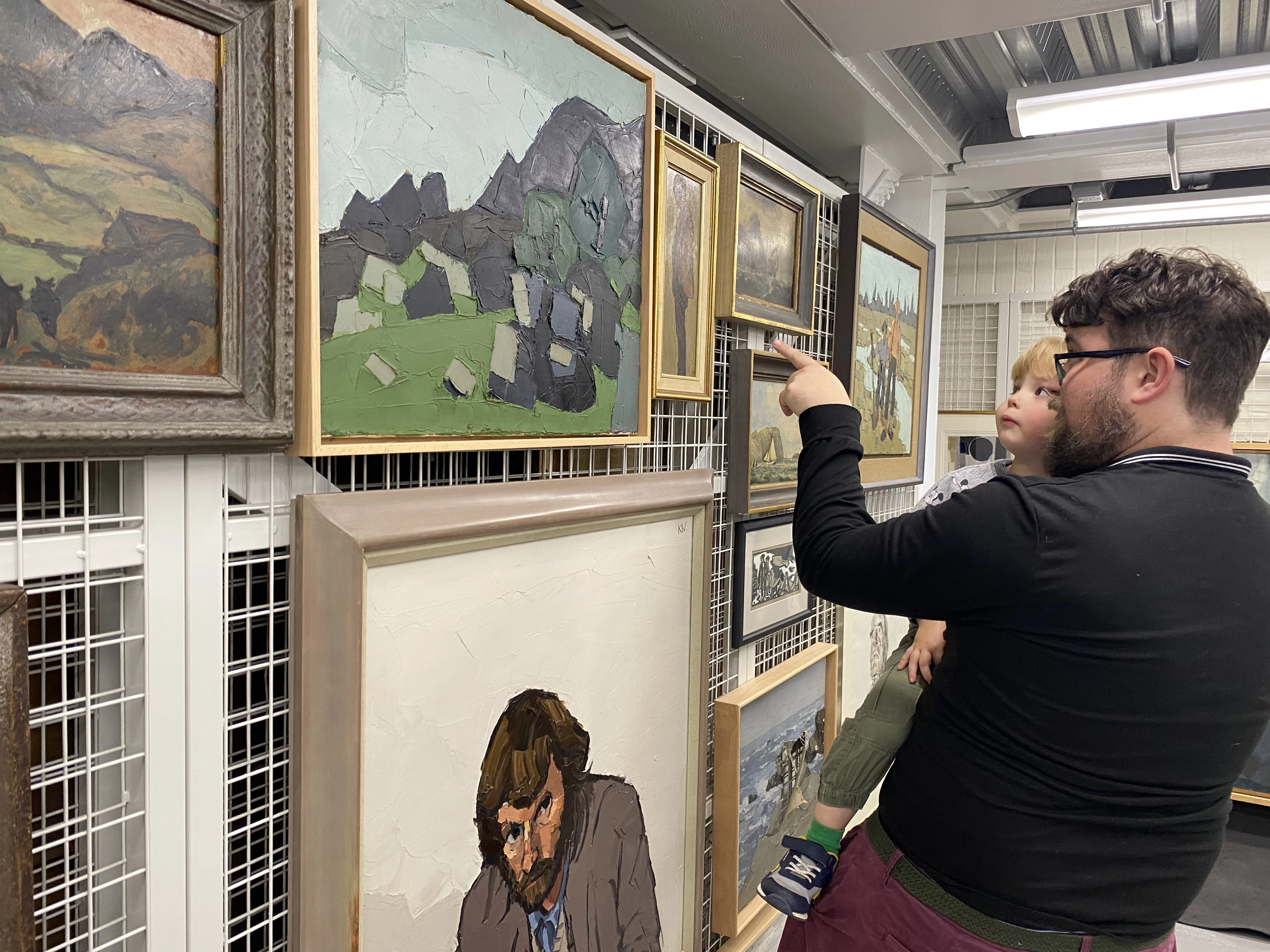 A man holds a child in his arms and points at a rack holding several artworks. They're both looking intently at the rack within an art store.