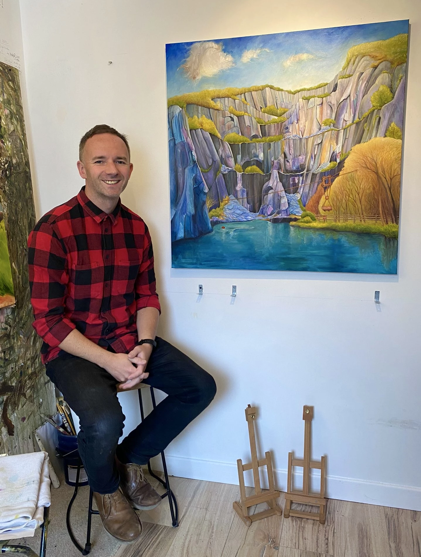 A man in a red and black check shirt sits on a high stool. To the right of him is a colourful painting of a slate quarry.