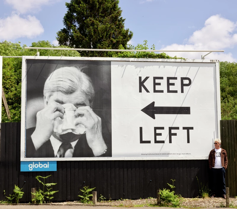 Billboard with a black and white photograph of Aneurin Bevan and 'Keep Left' in black capital letters with a black arrow pointing left. The artist stands to the right.