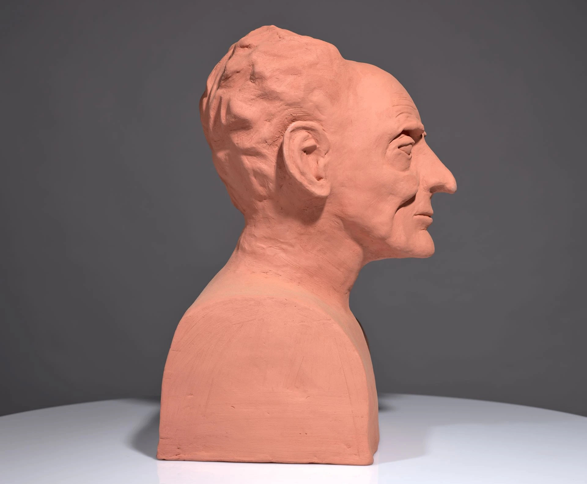 Bust of Saunders Lewis in orange clay. It sits on a grey background and a light grey base. Side profile as it faces to the right.
