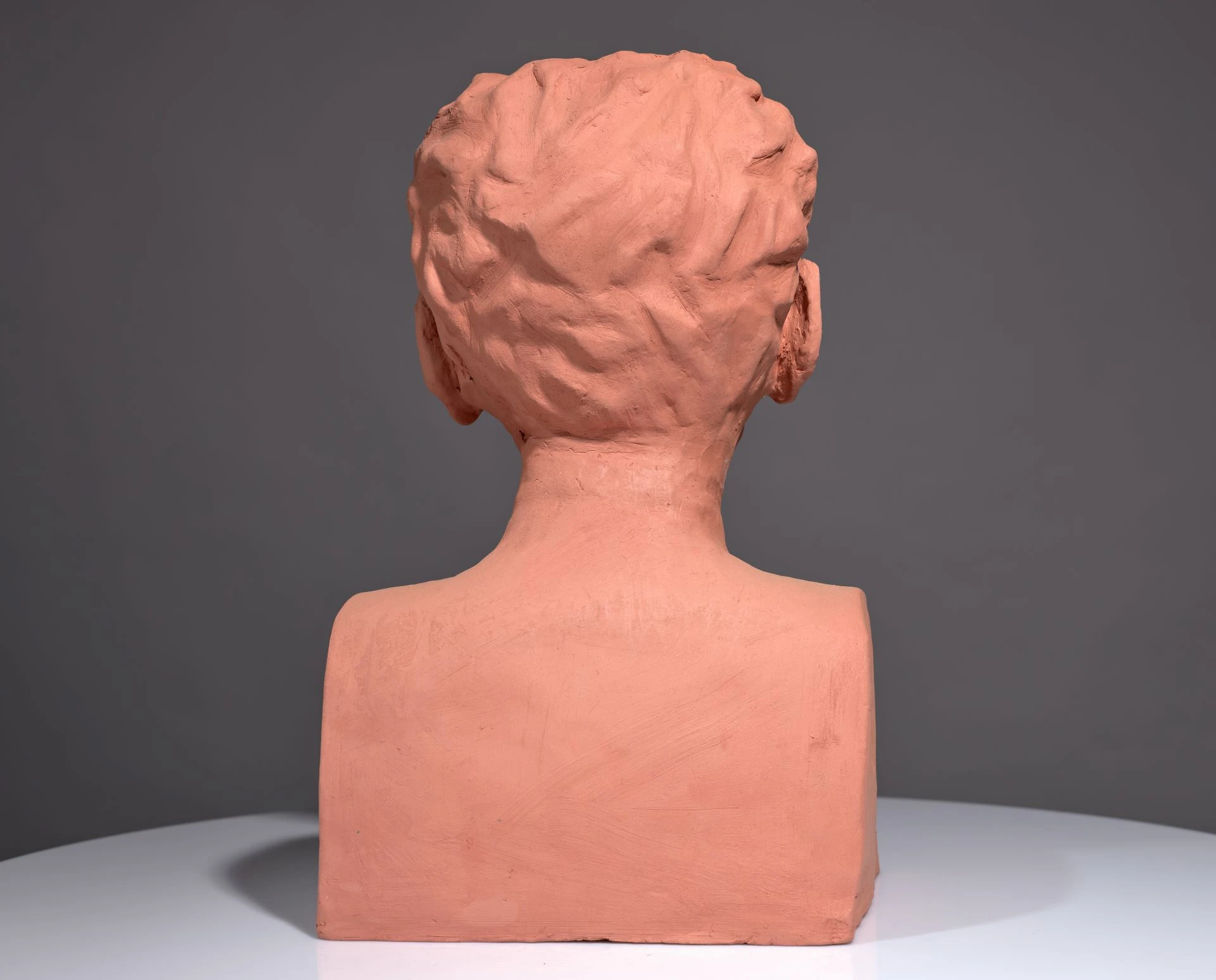 The back of a bust of Saunders Lewis in orange clay. It sits on a grey background and a light grey base