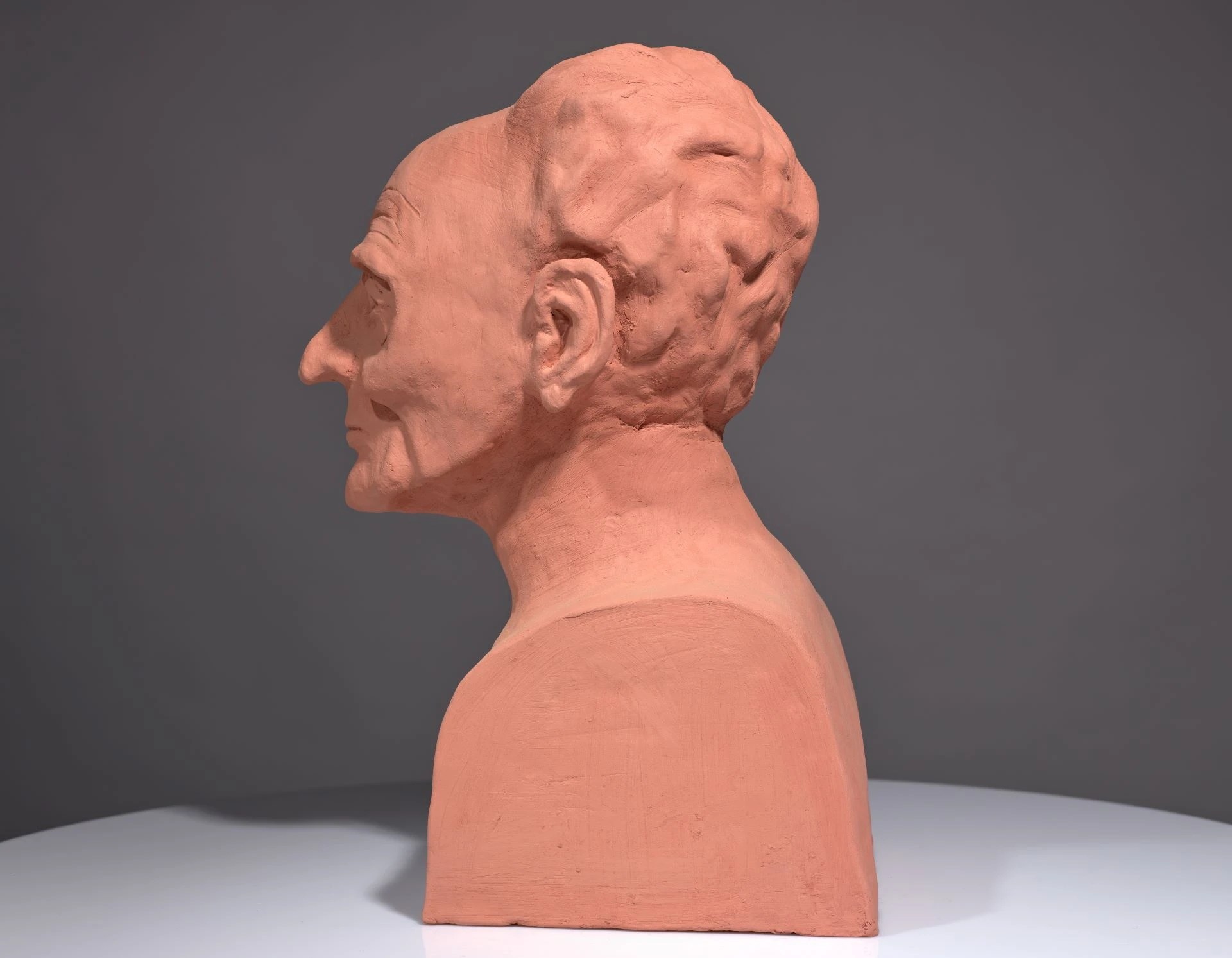 Bust of Saunders Lewis in orange clay. It sits on a grey background and a light grey base. The bust faces left.