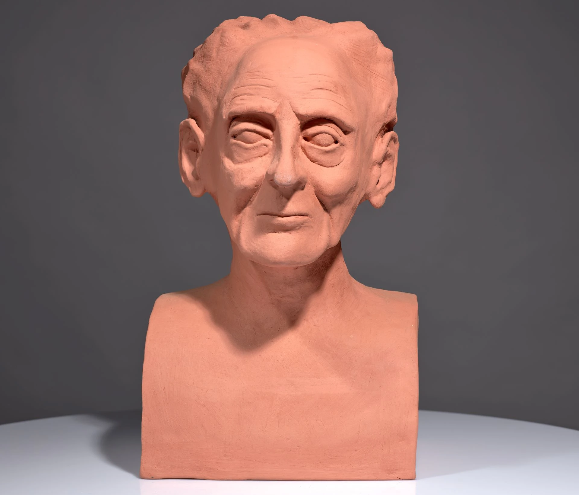 Bust of Saunders Lewis in orange clay. It sits on a grey background and a light grey base. It faces straight ahead.