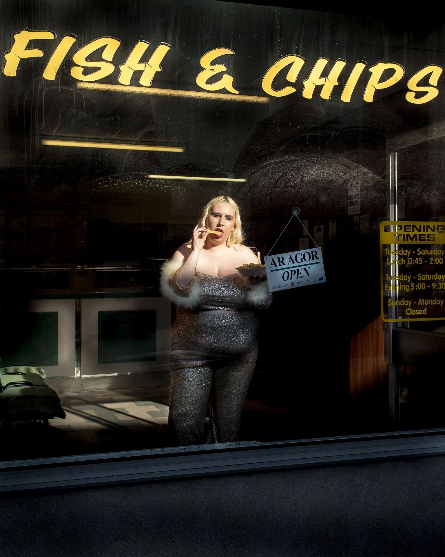 A blonde woman stands in the window of a fish and chip shop eating a tray of chips. She wears a gold sparkly jumpsuit.