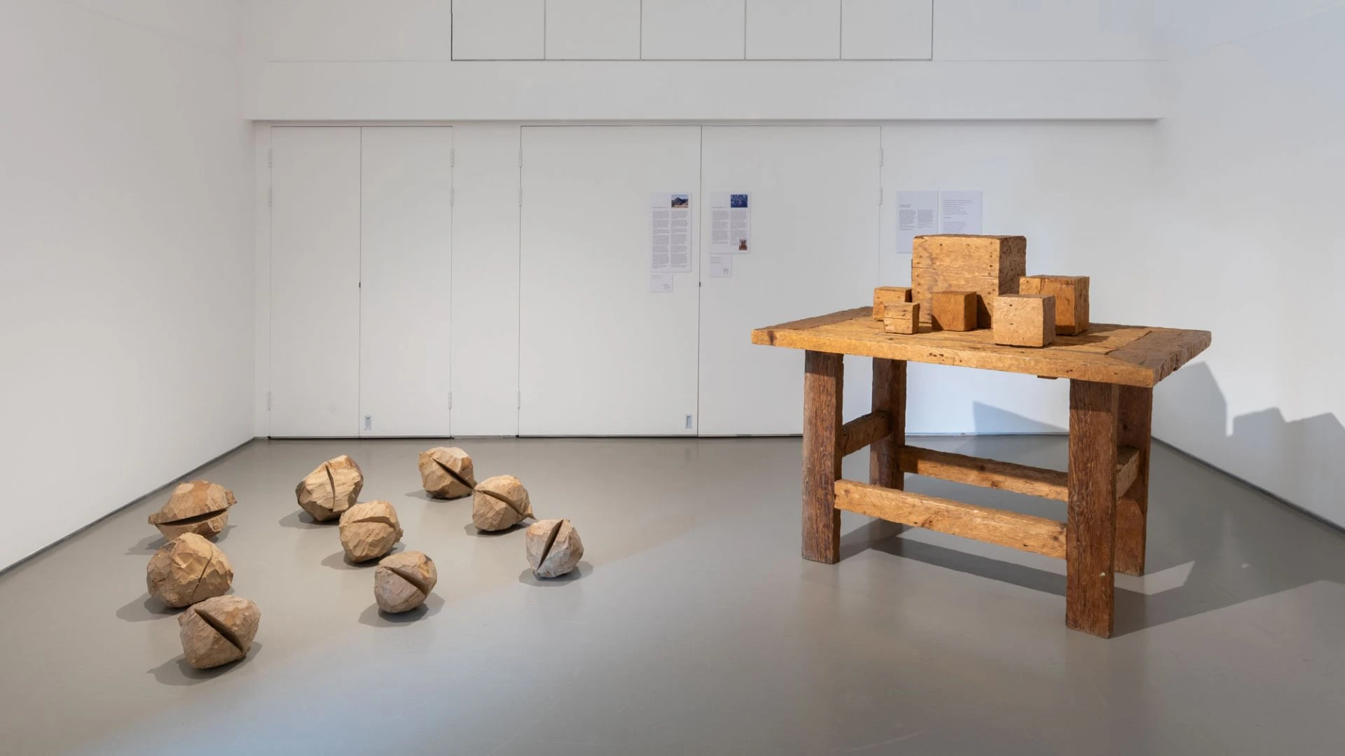 A white gallery space with a wooden table on the right with wooden blocks placed on it. On the floor on the left are 9 spheres of wood.