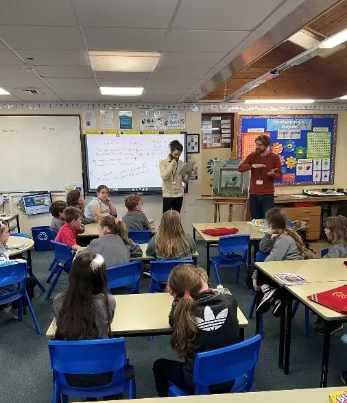 Two adults stand in front of a classroom of pupils.