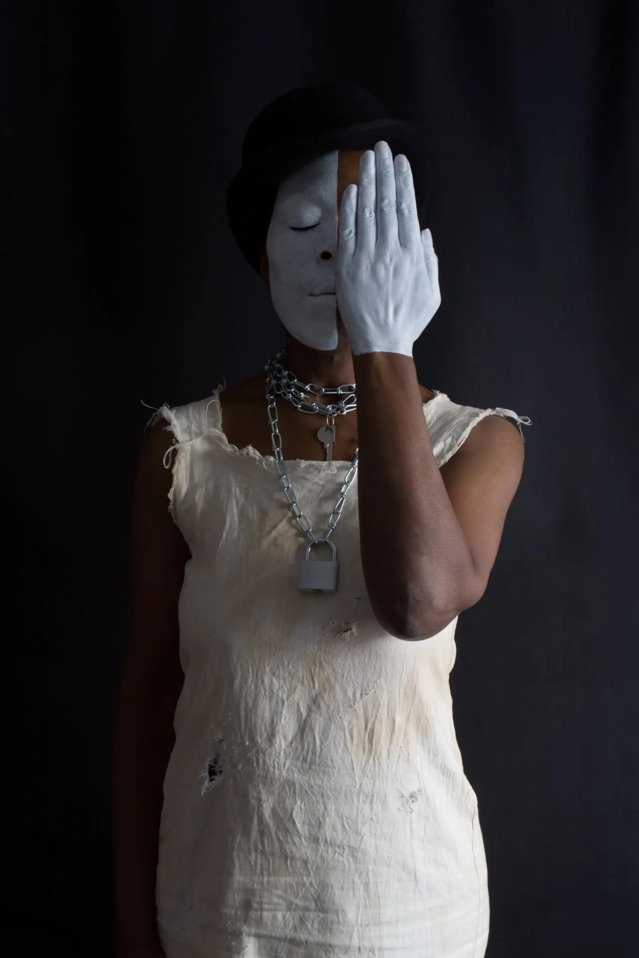A black woman with half her face covered in white facepaint. The other half of her face is hidden by her hand which is also covered in white paint. She wears a white dress with chains around her neck.